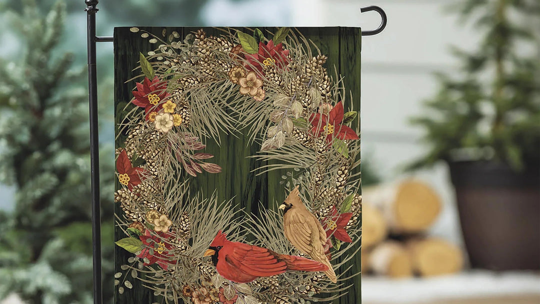 A decorative garden flag hanging from a black metal stand outdoors. The flag features a winter wreath design with red cardinals, evergreen branches, berries, and the text 'Hello WINTER' at the bottom.