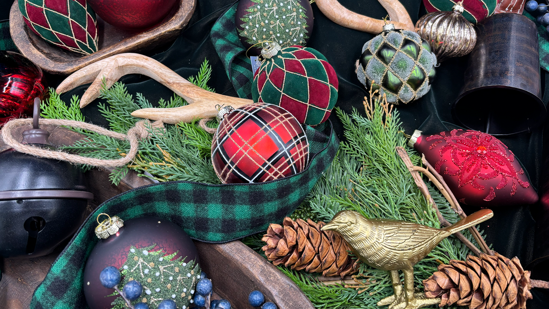 A styled holiday arrangement of Christmas ornaments, green plaid ribbon, pine branches, pinecones, blue berries, wooden antlers, bells, and a small gold bird ornament on a dark fabric background.