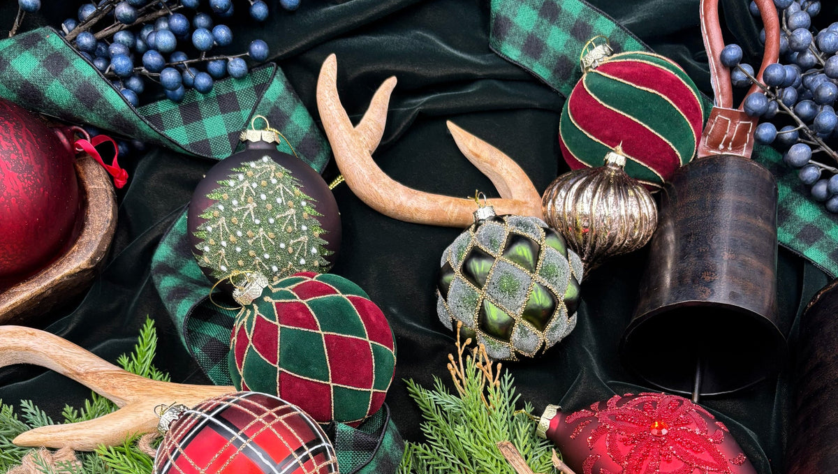 A styled holiday flat-lay featuring a collection of Christmas ornaments, including red and green plaid and striped baubles, a gold bird figurine, pinecones, green pine branches, blue berries, and plaid ribbons on a black fabric background.